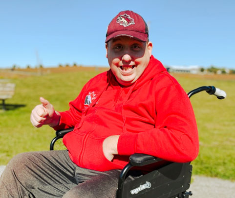 A smiling man in a wheelchair wearing a red shirt giving the thumbs up sign.