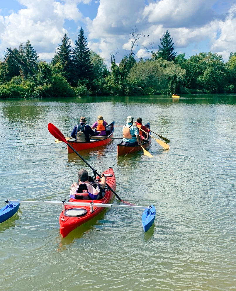 People padding two canoes and a Kayak on a lake with trees and a blue cloudy sky.