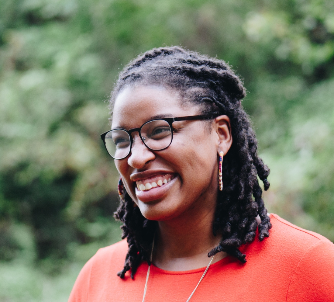 A smiling black woman with her hair in corn rows wearing an orange shirt