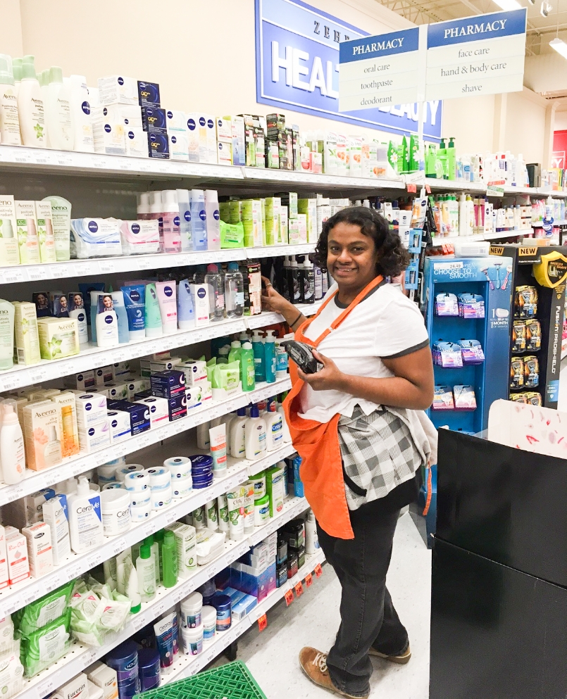 A smiling woman wearing an orange apron while stocking shelves in a pharmacy.