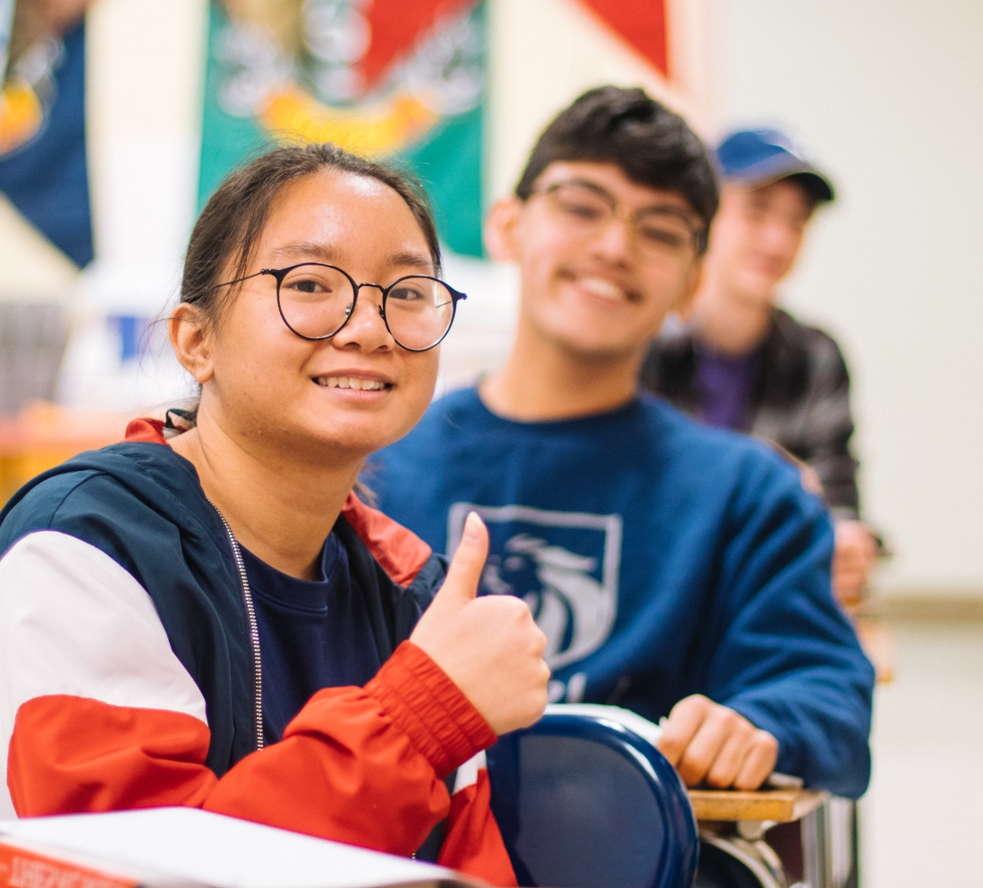 A young teen girl and 2 boys sitting in a row of desks. The girl is giving a thumbs up.