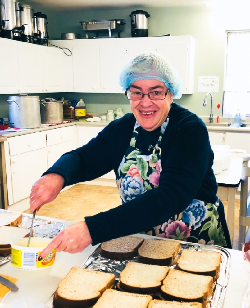 A smiling woman in a kitchen wearing a hair net while making sandwiches.