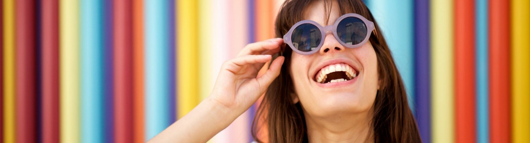 A smiling woman in purple sunglasses in front of a very colourful stripped wall.