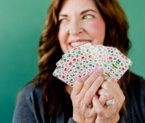 A smiling woman holding a handful of colourful playing cards up close to her face.