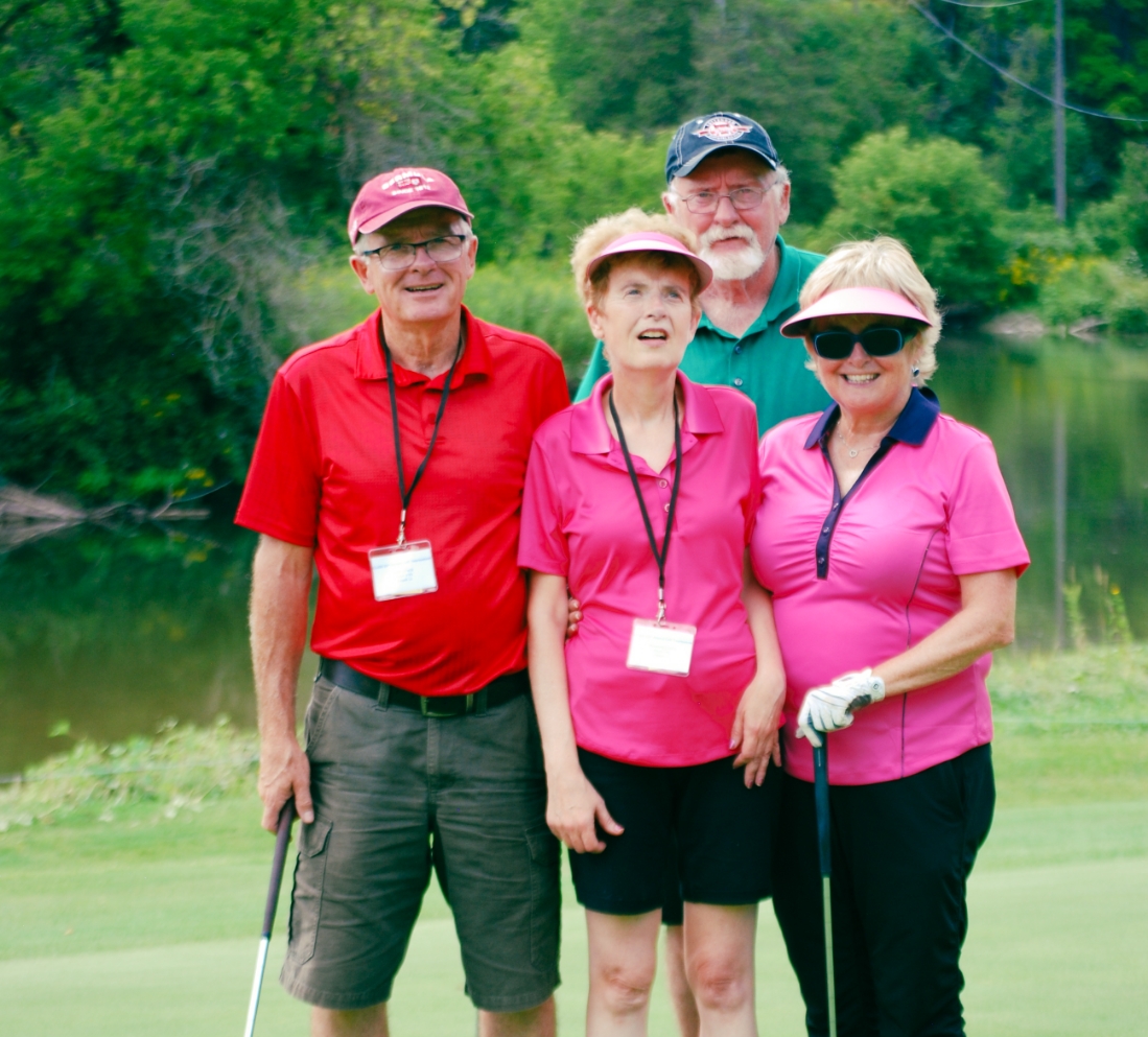 Two men and two woman wearing golf clothes and holding golf clubs. There is green trees and a pond behind them.