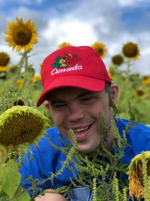 A young man smiling while surrounded by a field of sunflowers. There is a blue sky with white clouds in the background.