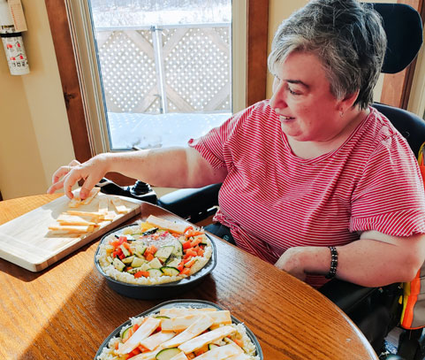 An older woman sitting in a wheelchair at a table adding cut cheese to her salad.