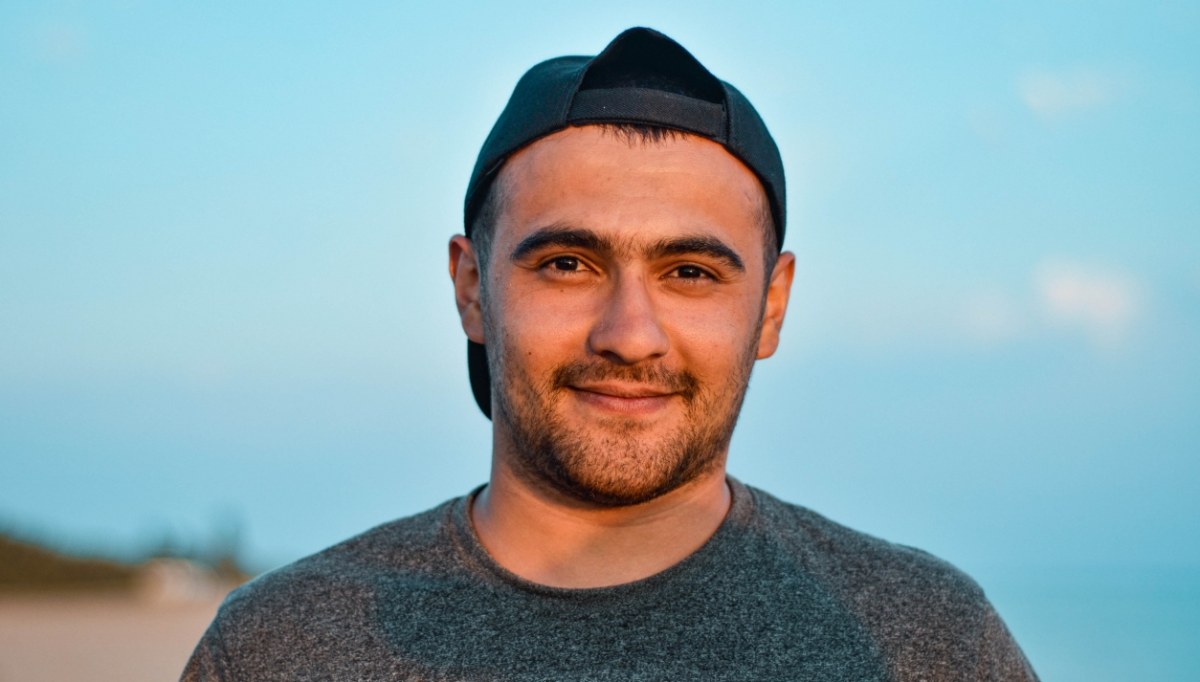 A young man with a short beard wearing a ball cap backwards with a nice blue sky in the background.