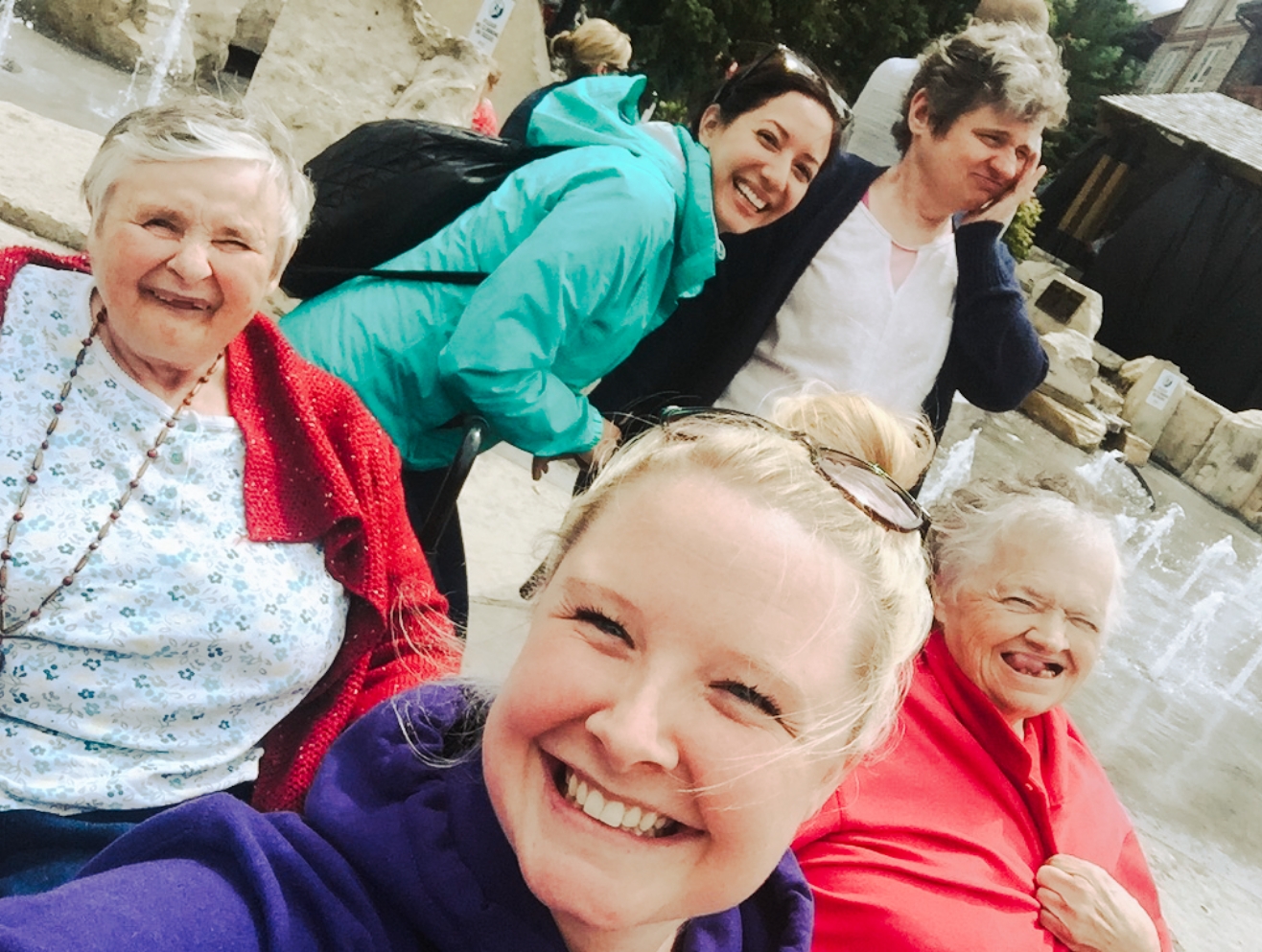 Five woman of varying ages laughing and smiling while taking a selfie with water fountains in the background.