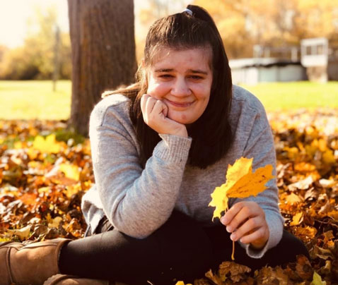 A young adult woman sitting in a pile of colourful leaves and holding a yellow leaf.