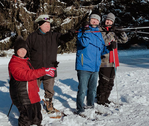 Five men of varying ages dressed in their winter clothes snowshoeing with evergreen trees behind them.