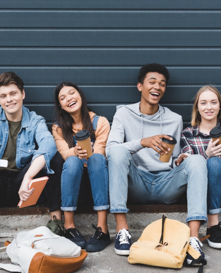 Four young teens sitting with their backs against a dark wall. There are backpacks at their feet.