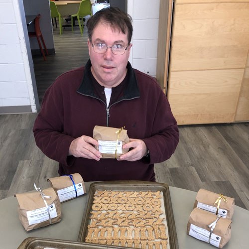 A man holding a bag of dog cookies with a tray of newly baked dog cookies on the table in front of him