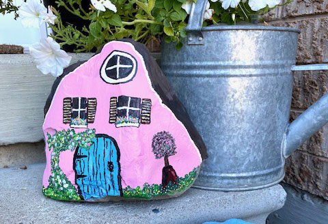 metal watering can with a rock painted like a pink house with shutters and a blue door