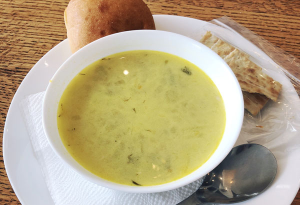 Close up of a bowl of yellow coloured soup on a plate with crackers, a bun and a spoon.